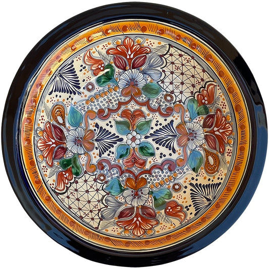 A round ceramic plate with a hand-painted Mediterranean style pattern, featuring orange, red, green, blue, and white floral motifs on a cream background, surrounded by a dark blue rim.