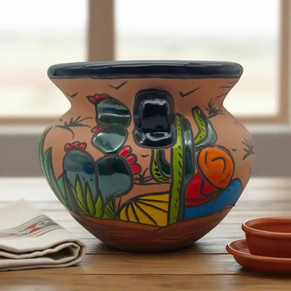 A hand-painted terracotta pot with a Southwestern cactus design, sitting on a wooden table with a folded cloth napkin and a small matching terracotta bowl in the foreground.